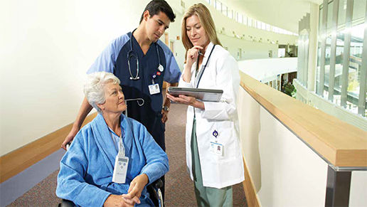 An elderly woman in a wheelchair, wearing a blue robe and oxygen cannula, being attended to by a male nurse a female doctor. 