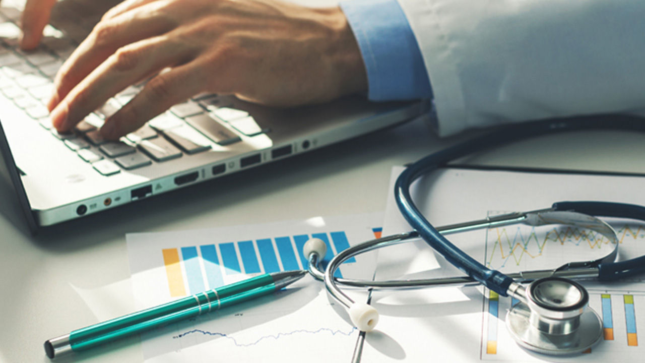 healthcare provider typing on a laptop with a stethoscope and papers next to it