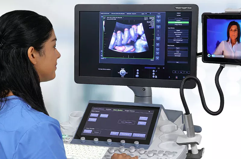 Nurse at a computer looking at imaging results on one screen and talking to a doctor on the other screen
