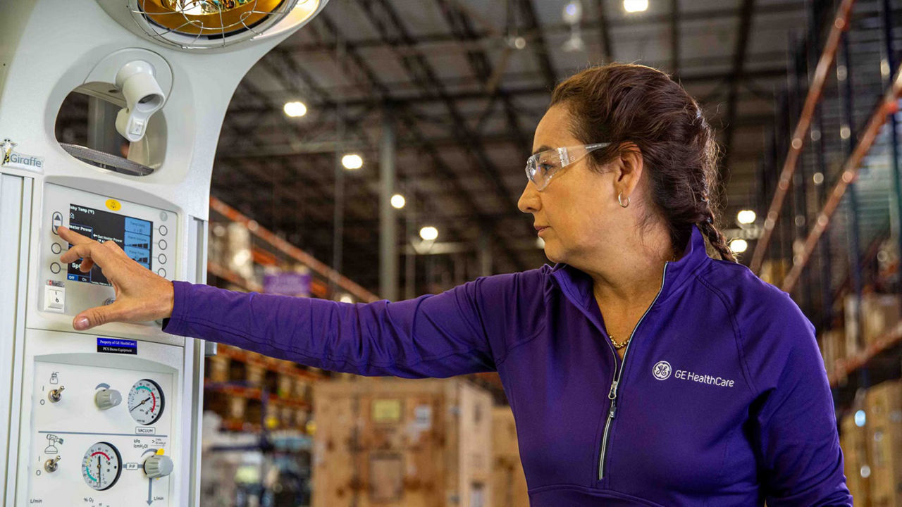 GE HealthCare technician wearing protective eye gear while adjusting a setting on a piece of critical care equipment in a warehouse.