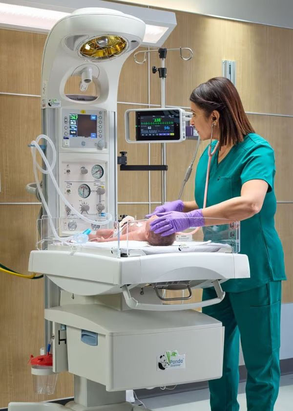 Clinician adjusting a setting on a Panda Warmer while an infant rests in a hospital bassinet.