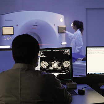 Image of a clinician monitoring CT scans on two monitors while another clinician uses a CT scan machine in the background.