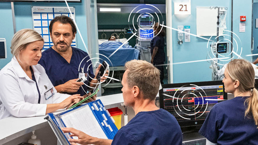 four hospital staff talking at a desk with a patient in a bed in the background