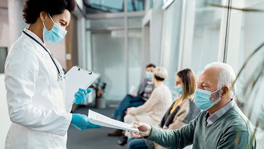 Healthcare professional wearing a mask handing paperwork to an elderly patient in a medical waiting area.