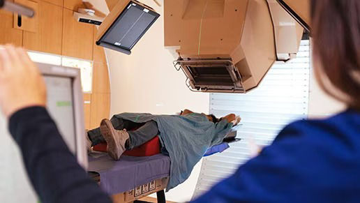 Patient positioned on a treatment table receiving radiation therapy, with a clinician operating the treatment system from the control area.