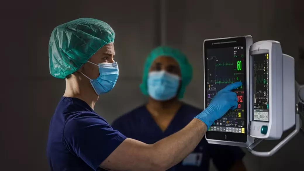 A clinician wearing a hair net, a face mask and gloves using a touchscreen monitor device. Another clinician is in the background.