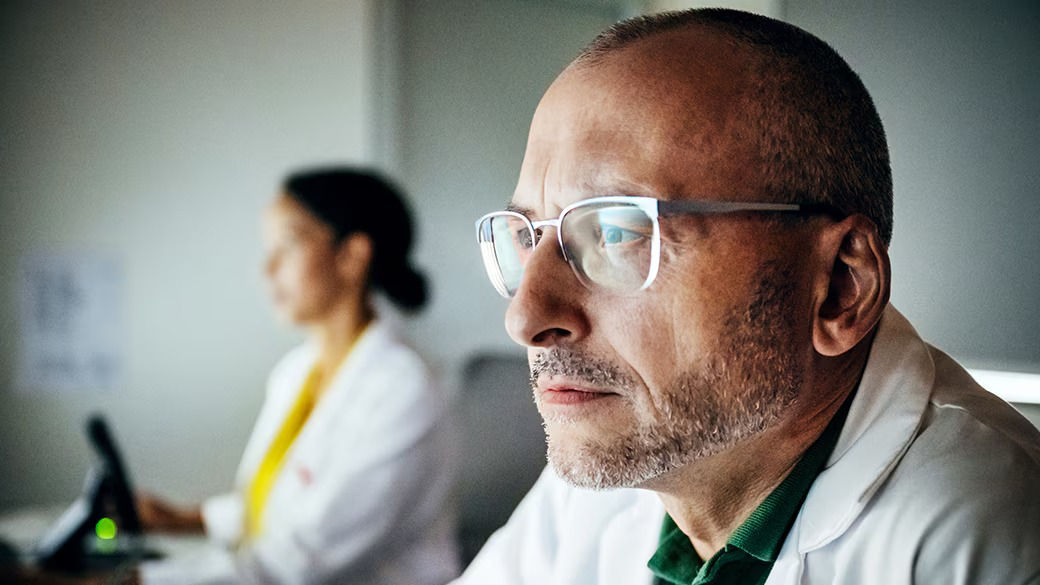 A man in a white coat wearing glasses. A woman in a white coat using is using a computer behind the man.