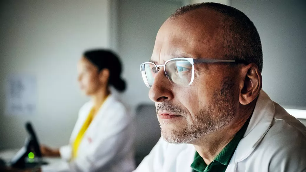 A man in a white coat wearing glasses. A woman in a white coat using is using a computer behind the man.