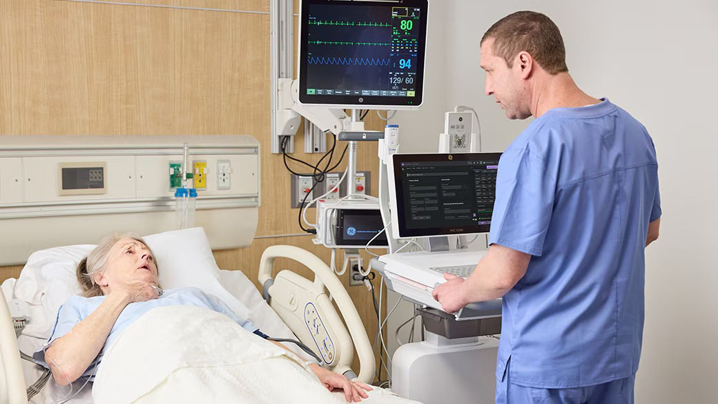 A clinician using a computer while talking to a patient who is lying in a hospital bed.