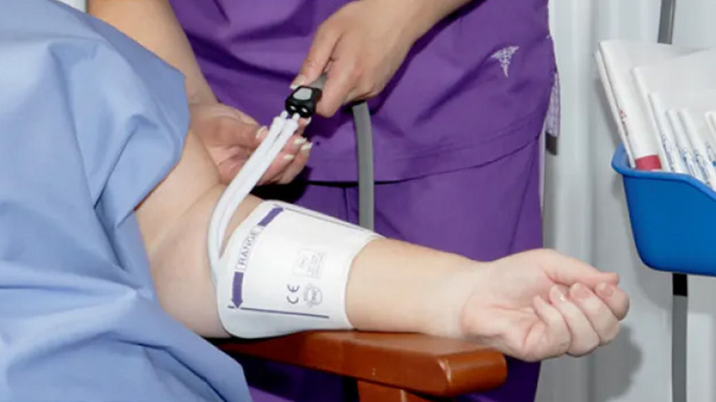 Person sitting in chair wearing hospital gown with another person in purple scrubs taking their blood pressure