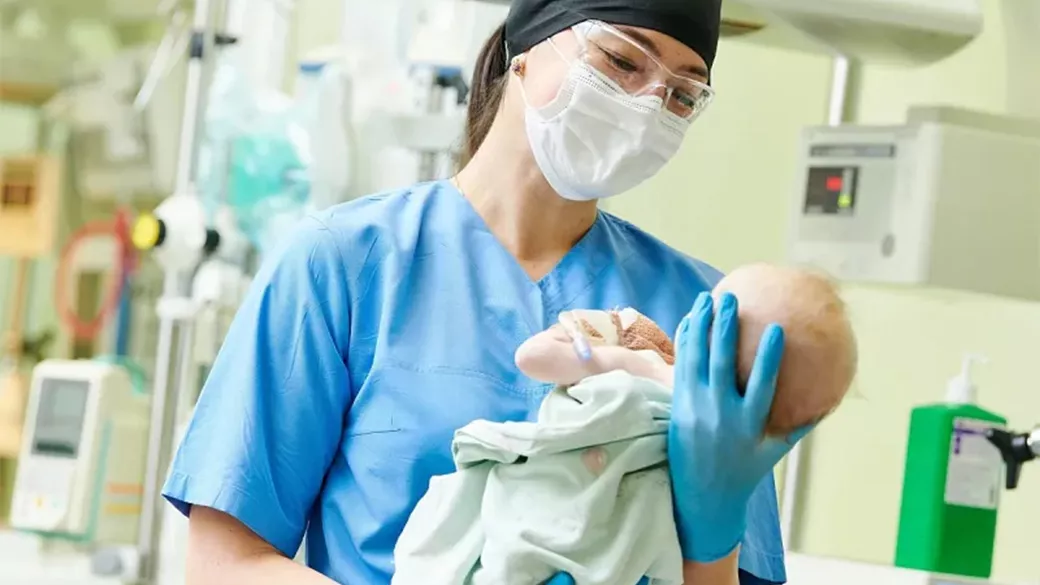 A clinician wearing a face mask and gloves while cradling a newborn baby in a delivery room.