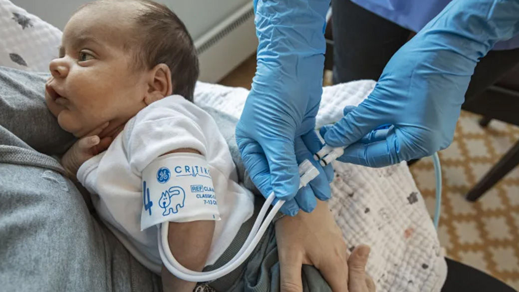 Baby being held by one person in a hospital bed. Another person is taking the baby's blood pressure and wearing blue gloves