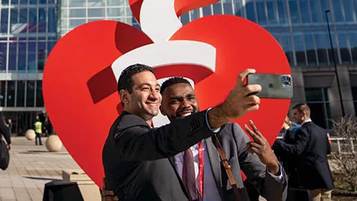 Image of two men talking a picture with a cellphone in front of an AHA sign.