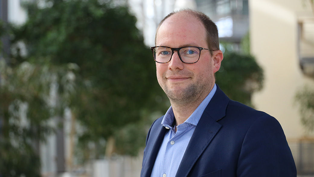 Image of a man in a blue suit and glasses standing in front of a building and trees smiling at the camera.