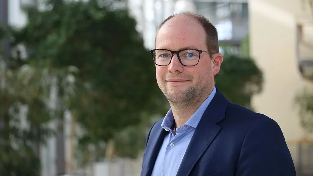 Image of a man in a blue suit and glasses standing in front of a building and trees smiling at the camera.