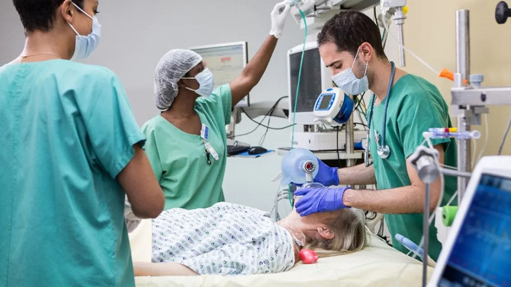 Clinician wearing a facemask administering anesthesia to a patient lying on an operating table. A second clinician wearing a scrub cap and facemask engaging with a monitor in the background. A third clinician with a facemask standing in front of the operating table.