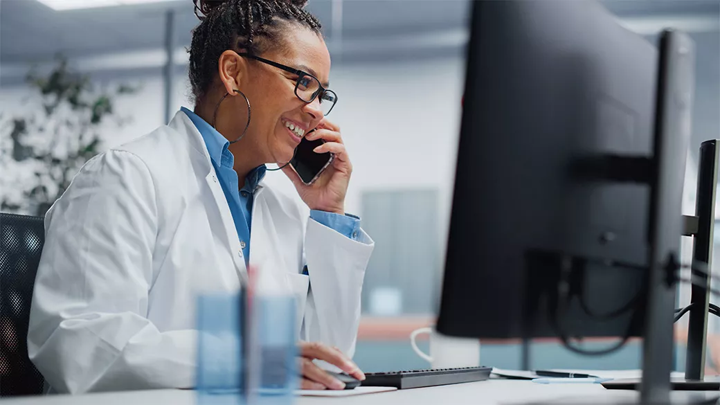 Doctor sitting at a desk in front of a desktop monitor and holding a phone to her ear
