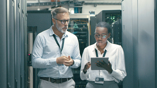 two technicians walk down aisle of servers in discussion while reviewing tablet