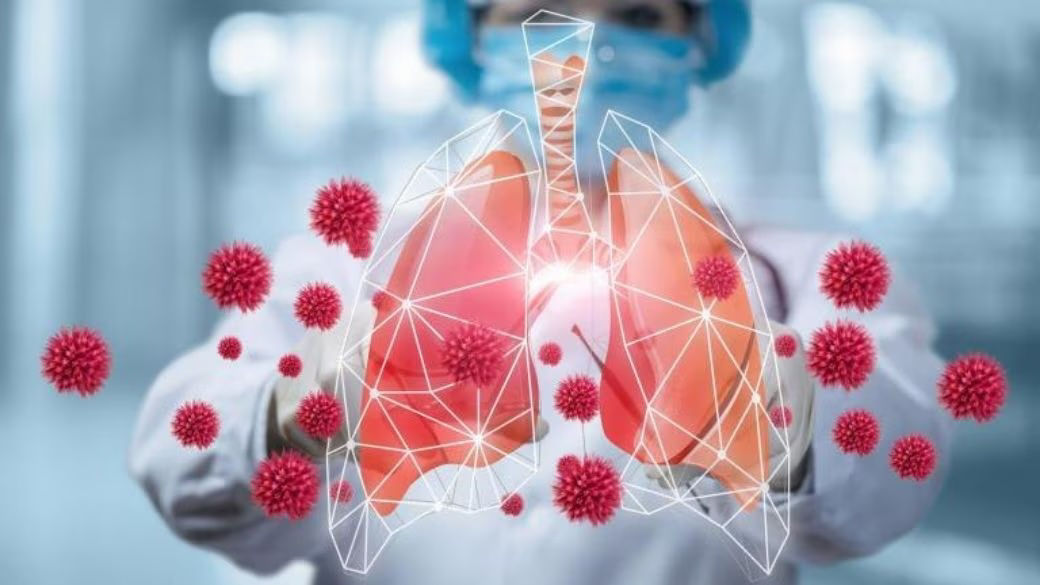 In the background, a healthcare worker is shown reaching her arms out in front of her. She is wearing full PPE, including a surgical mask, a white coat, gloves, and a disposable surgical bouffant caps. In front of her out stretched arms is a pictogram of a pair of lungs, trachea, and large particles. 