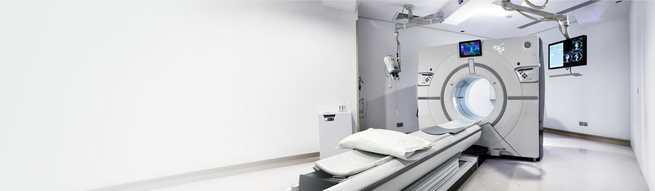 A modern medical imaging scanner stands in a bright, white hospital room. The machine features a circular opening with a patient table extending toward it, prepared with cushions and sheets. Overhead equipment and monitors display imaging information, emphasizing advanced diagnostic technology. The clean, minimalist environment suggests a sterile, professional healthcare setting.