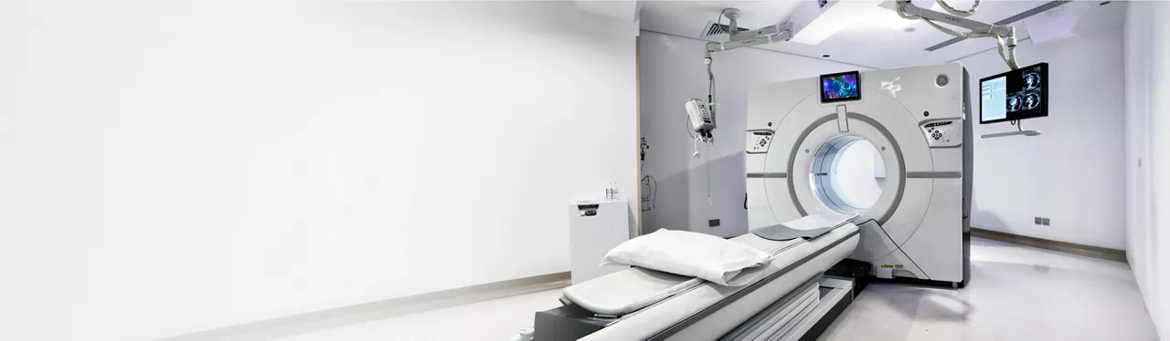 A modern medical imaging scanner stands in a bright, white hospital room. The machine features a circular opening with a patient table extending toward it, prepared with cushions and sheets. Overhead equipment and monitors display imaging information, emphasizing advanced diagnostic technology. The clean, minimalist environment suggests a sterile, professional healthcare setting.
