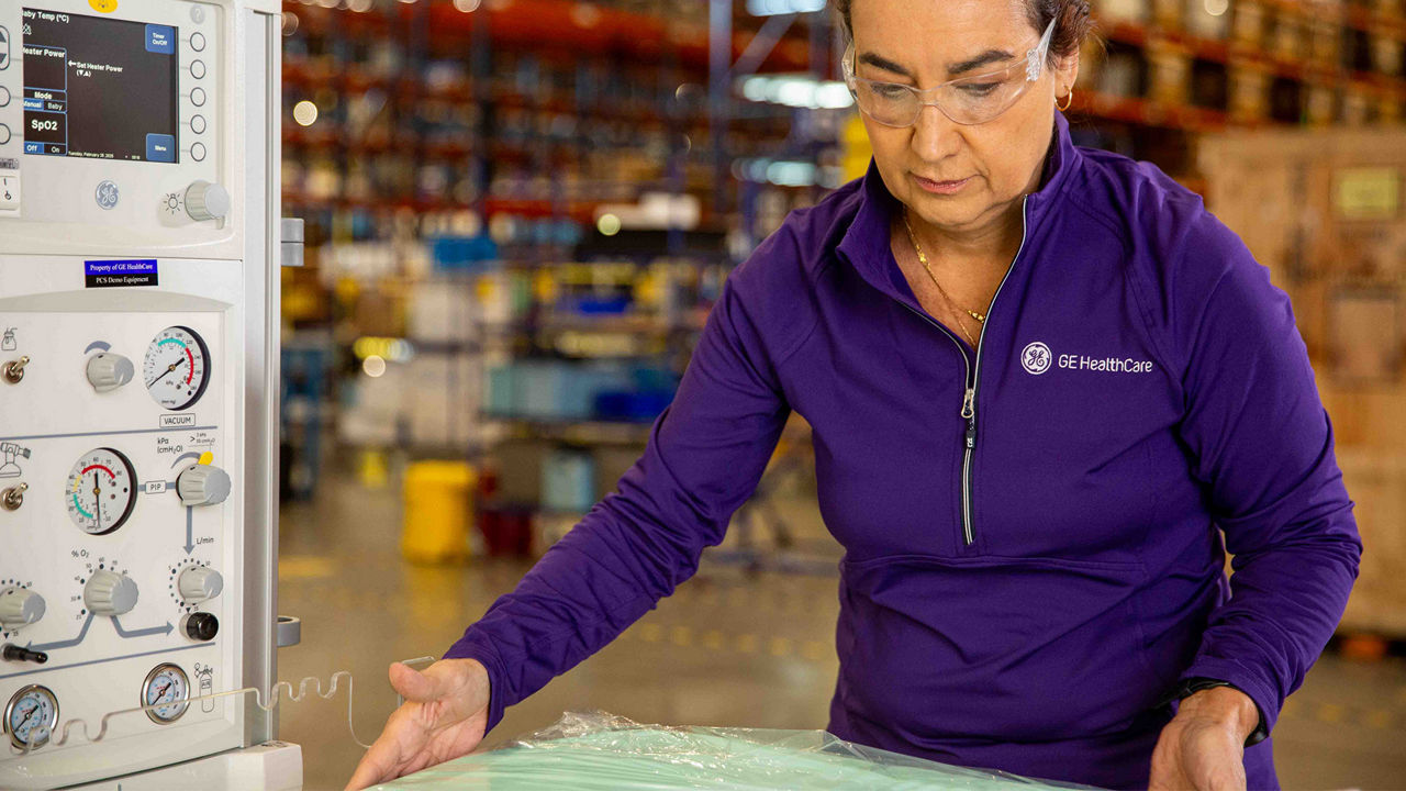 GE HealthCare technician wearing protective eye gear while adjusting a setting on a piece of critical care equipment in a warehouse.