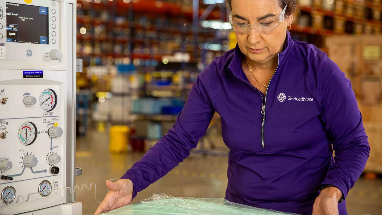 GE HealthCare technician wearing protective eye gear while adjusting a setting on a piece of critical care equipment in a warehouse.