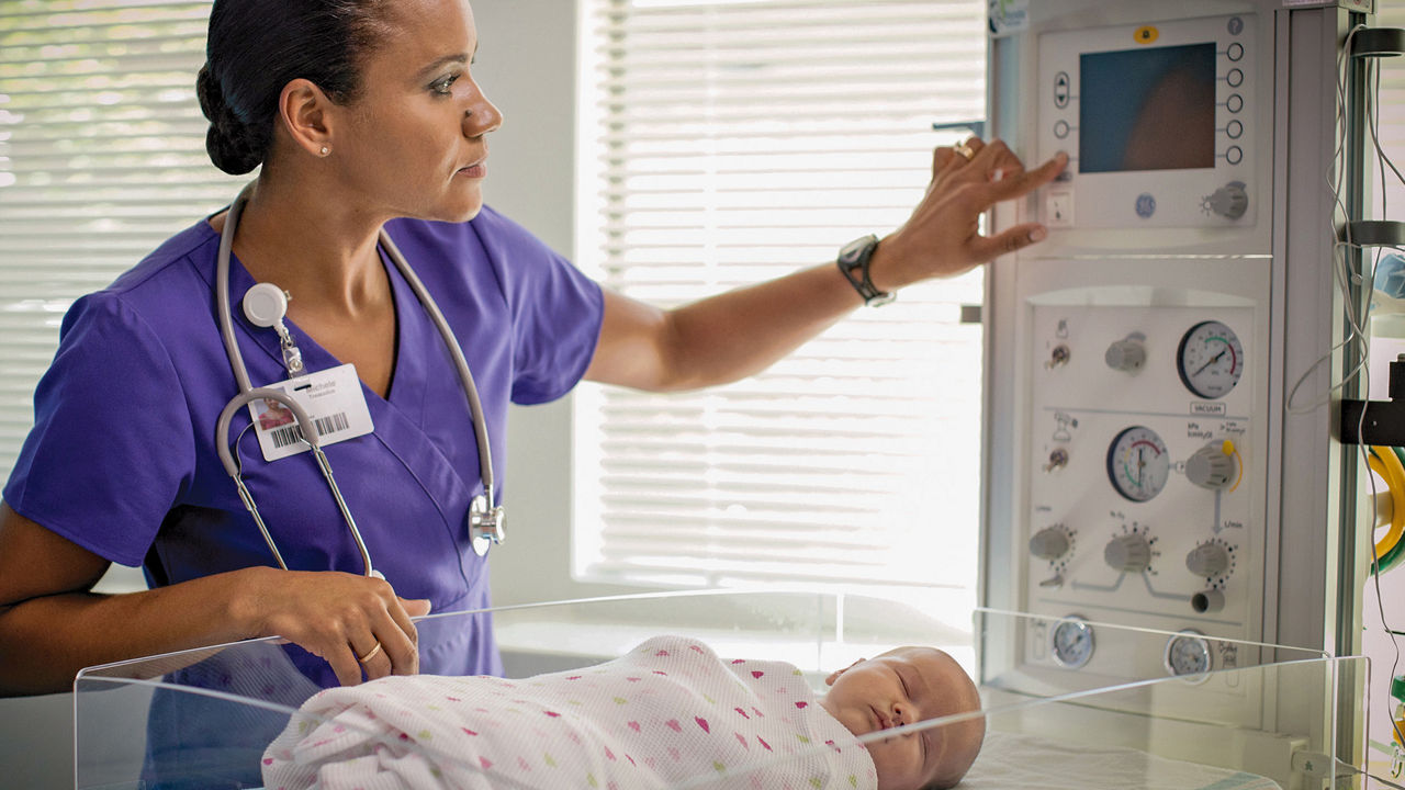 Clinician adjusting a setting on a Panda Warmer while an infant rests in a hospital bassinet.