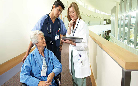An elderly woman in a wheelchair, wearing a blue robe and oxygen cannula, being attended to by a male nurse a female doctor.