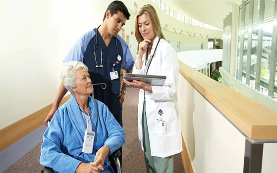 An elderly woman in a wheelchair, wearing a blue robe and oxygen cannula, being attended to by a male nurse a female doctor.