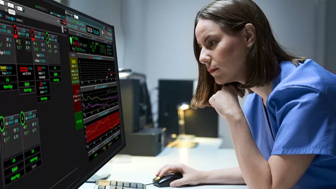 woman in blue scrubs stares at analytics on two monitors