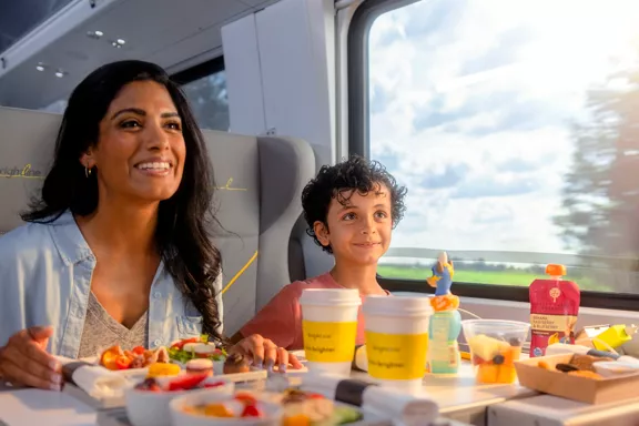 A mother and young child seated at a table aboard a Brightline train with food set out to eat