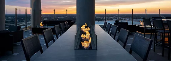 A contemporary rooftop terrace features a long rectangular fire pit centered on a sleek stone table. Dark chairs and bar stools line both sides, creating an inviting outdoor seating area. Glass railings frame the perimeter, opening to a wide cityscape view. A colorful sunset sky adds warm tones that complement the flickering flames and modern architectural elements.