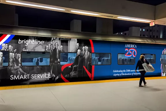 A woman walking down a train platform past a Brightline car featuring an "America 250" wrap.
