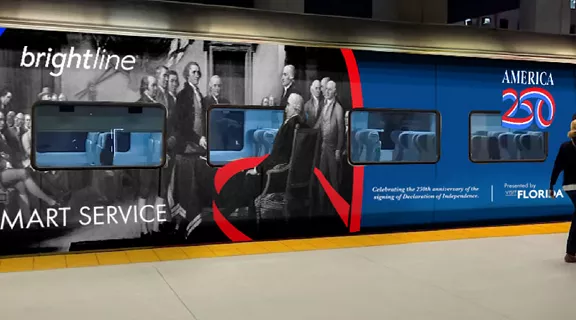 A woman walking down a train platform past a Brightline car featuring an "America 250" wrap.