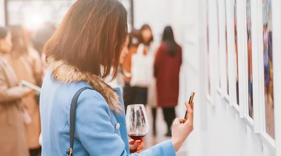 A woman at an art gallery holding a glass of wine and taking a picture of a painting with her phone.
