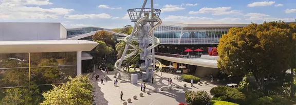 An open-air plaza features a tall, futuristic metal sculpture with looping structures at its center. People walk and gather around the installation, creating a lively public space atmosphere. Surrounding buildings with large glass windows and shaded walkways frame the scene, while trees and landscaped greenery soften the architecture. Bright daylight and a blue sky with scattered clouds give the image a vibrant, welcoming feel.