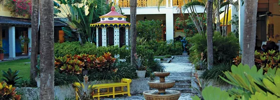 A vibrant courtyard garden features lush tropical plants surrounding a small multi-tiered stone fountain. A colorful gazebo with a red roof and decorative columns stands in the background, framed by palm trees and flowering shrubs. Yellow benches and bright building facades add pops of color to the tranquil outdoor setting. The scene suggests a peaceful, inviting space for relaxation in a warm climate.