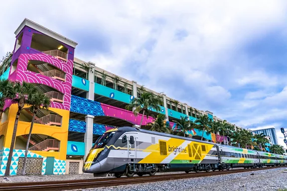 Brightline's Bright Green train on railroad tracks in front of colorful building