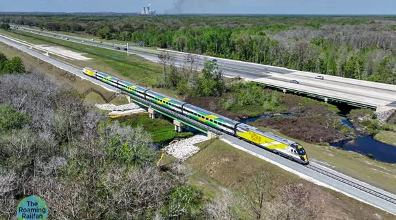 Brightline train passing through econ river bridge, overlayed with "The Roaming Railfan" globe icon in the bottom left corner