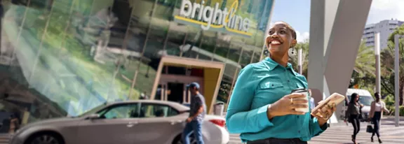 A smiling woman outside a Brightline station with a coffee in one hand and a smart phone in the other