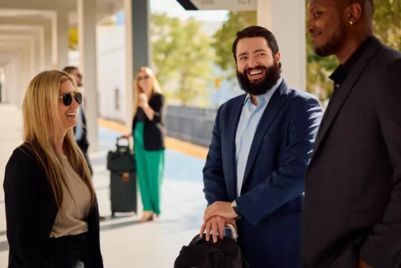 A group of men and women in business attire laughing and smiling while standing outside at a Brightline train station.