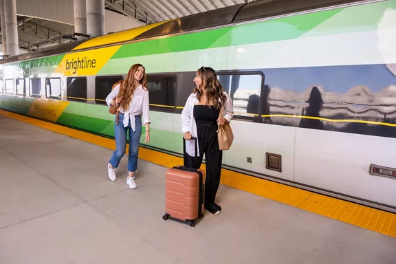 Two women carrying their luggage and smiling as they board a Brightline train.