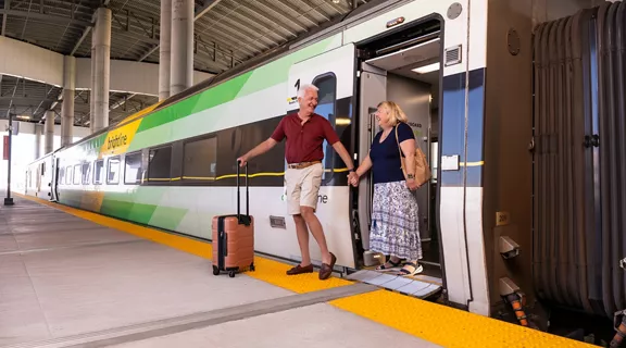 An older man and woman stepping off of a Brightline train with smiles on their faces.