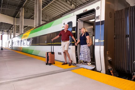 An older man and woman stepping off of a Brightline train with smiles on their faces.