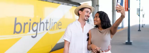 A man and woman posing for a selfie with a Brightline train car in the background.
