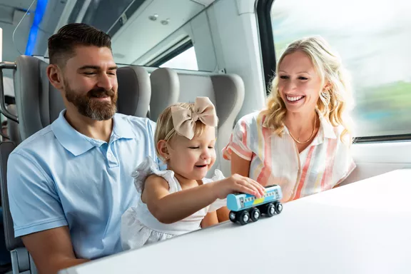 Two parents riding on a Brightline train with their young daughter, who is playing with a toy train
