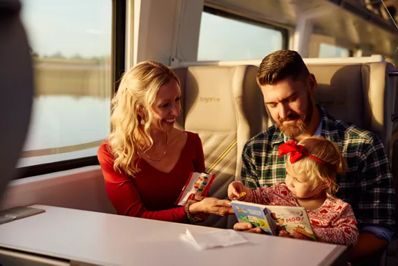 Parents reading a book to their young daughter while taking a ride on a Brightline train. 