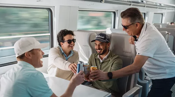 Four male friends dressed in golf attire, smiling and toasting with beers while riding on a Brightline train.