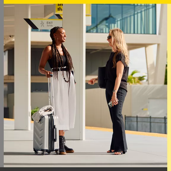 Two women stand and chat at an outdoor transit station, one holding a rolling suitcase and the other carrying a laptop and smartphone. 